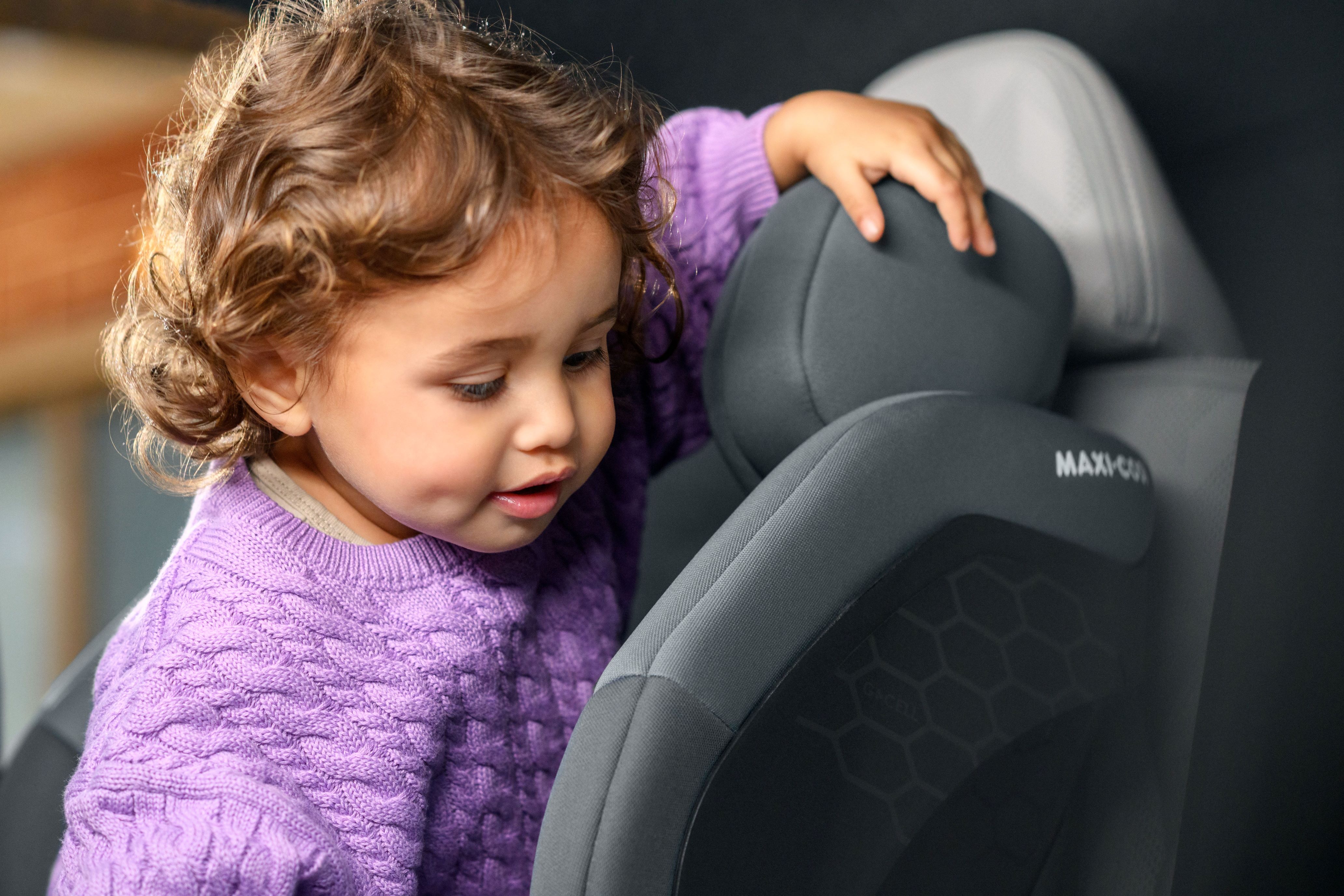 Kleines Mädchen im lila Pullover klettert neugierig auf einen Maxi-Cosi Kindersitz im Auto.
